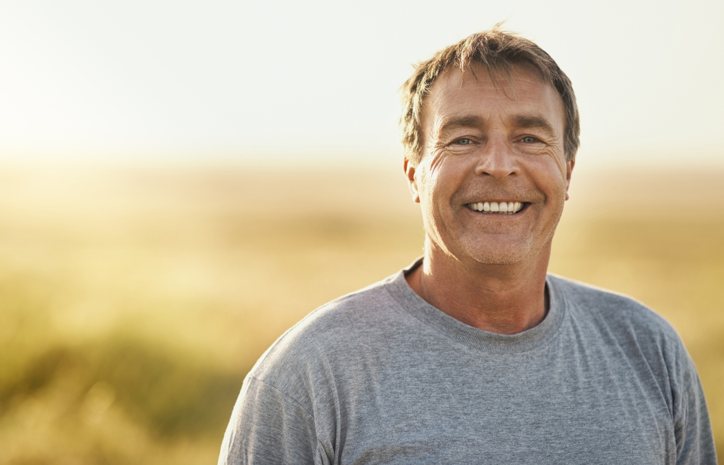 Middle-aged man smiling in a gray t-shirt, standing outdoors with a blurred natural background.