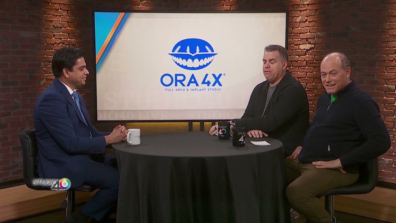 Three men sit around a round table on a TV studio set with a screen displaying the ORA 4X logo in the background.