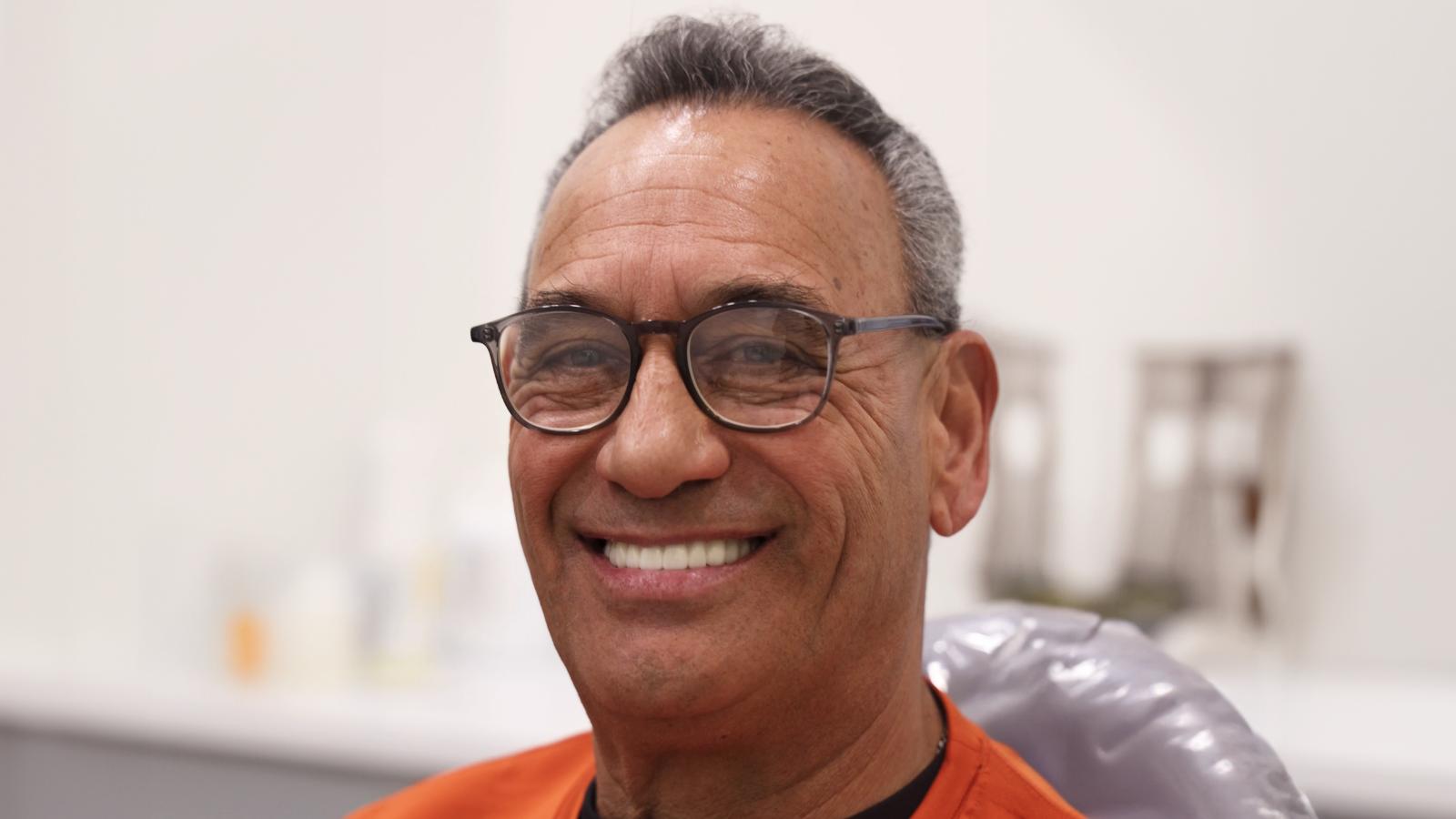 A middle-aged man with glasses and gray hair smiles while sitting in a chair, wearing an orange shirt in a brightly lit room.