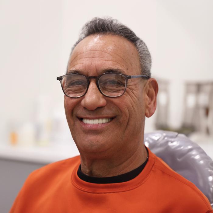 A man with short gray hair, wearing glasses and an orange sweater, sits in a chair and smiles at the camera in a brightly lit room.