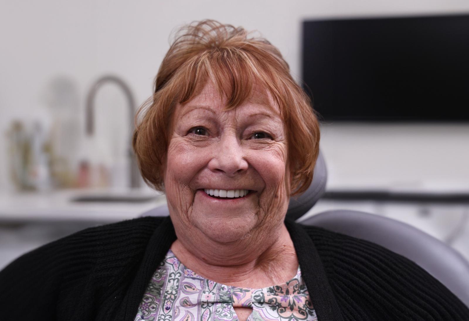 An older woman with short, reddish hair smiles while sitting in a dental chair in a modern clinic setting.