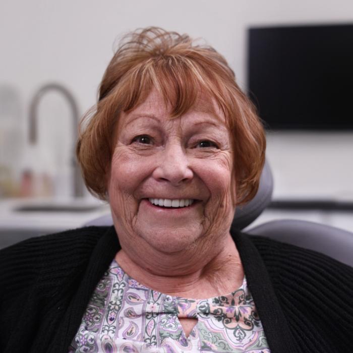 Older woman with short, light brown hair sits in a dental chair, smiling, wearing a patterned top and black cardigan. A sink and monitor are visible in the background.