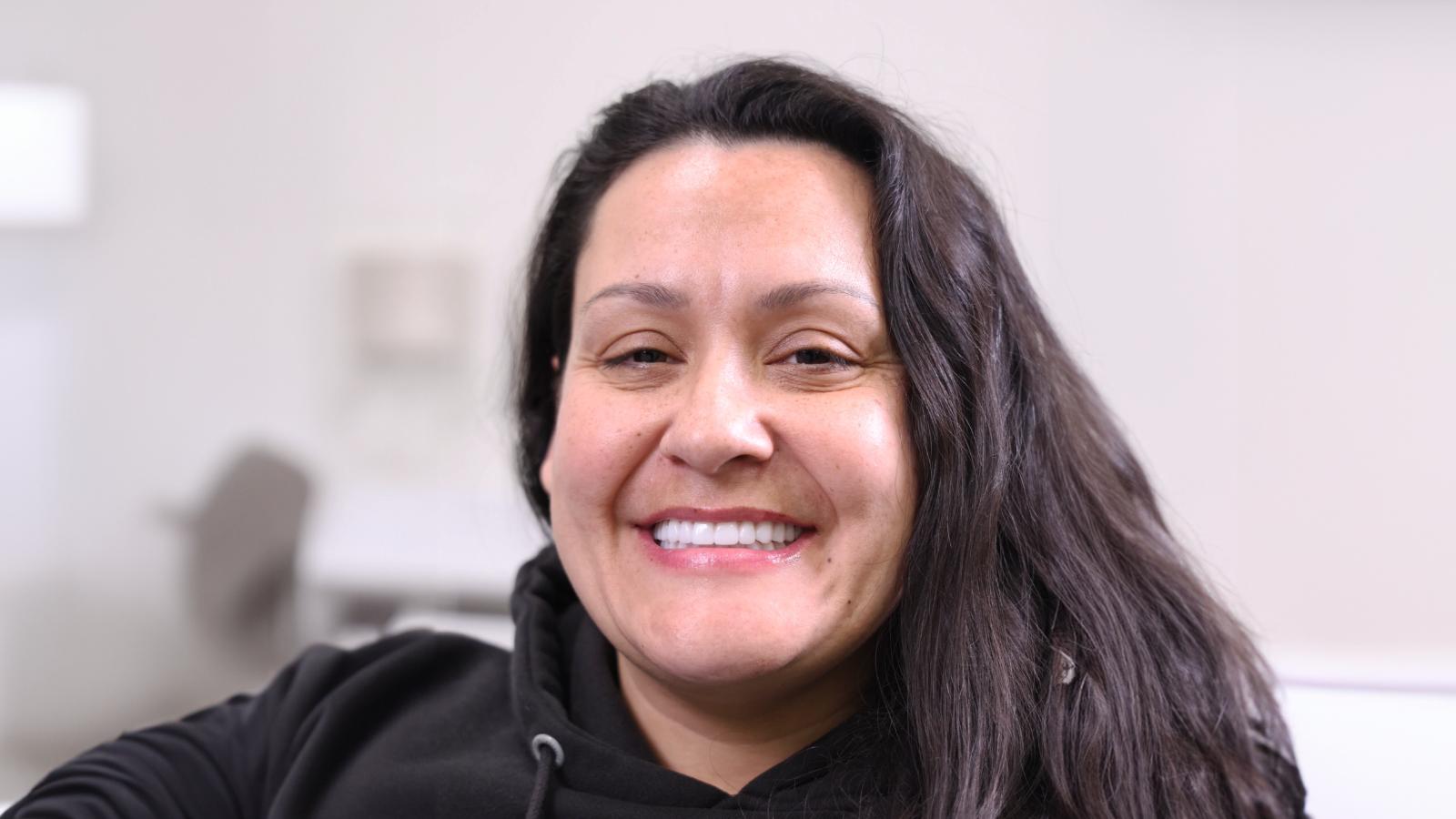 A woman with long dark hair wearing a black hoodie is smiling at the camera in a softly lit indoor setting.