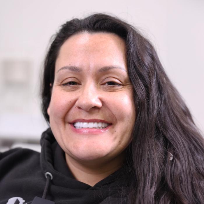 A woman with long dark hair smiles at the camera, wearing a black hoodie. The background is out of focus and neutral-colored.