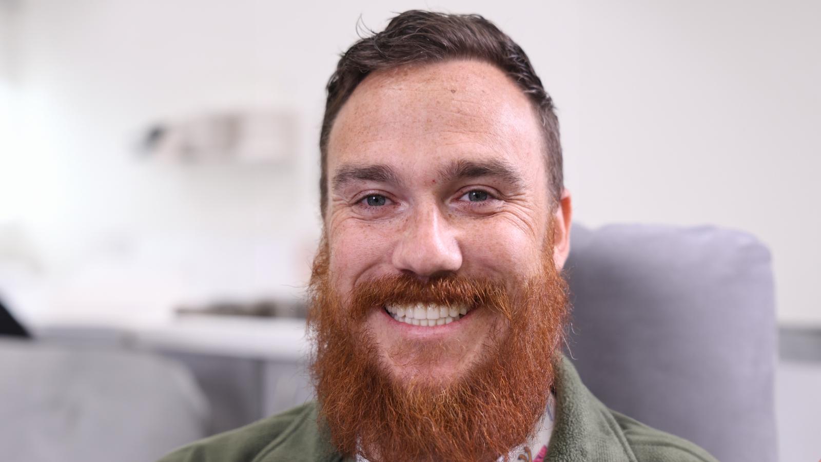 A man with a reddish-brown beard and mustache is smiling at the camera in a bright indoor setting.