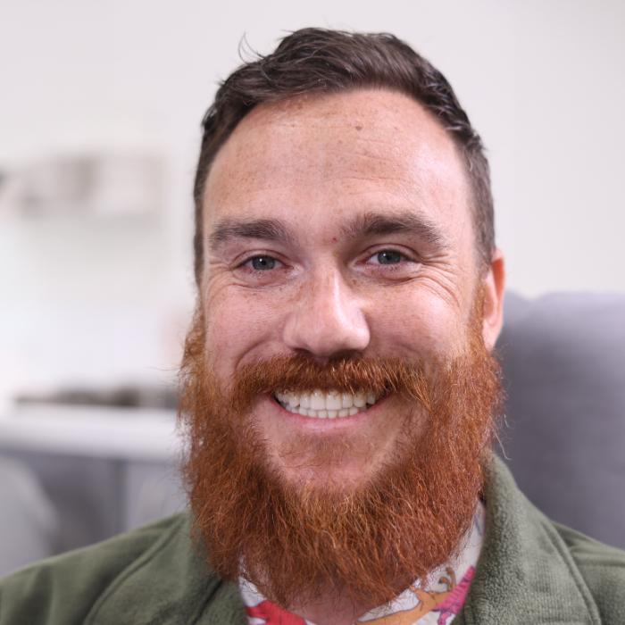 A man with a full red beard and mustache smiles at the camera, wearing a green jacket and a colorful shirt. The background is softly out of focus.