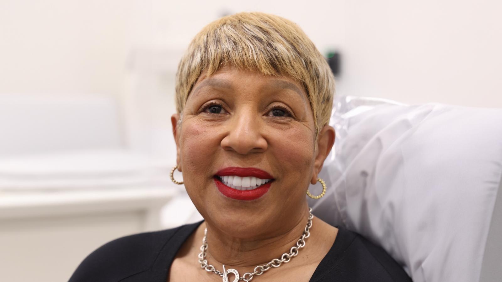A woman with short blonde hair, hoop earrings, and a necklace is sitting in a clinical setting, smiling at the camera.