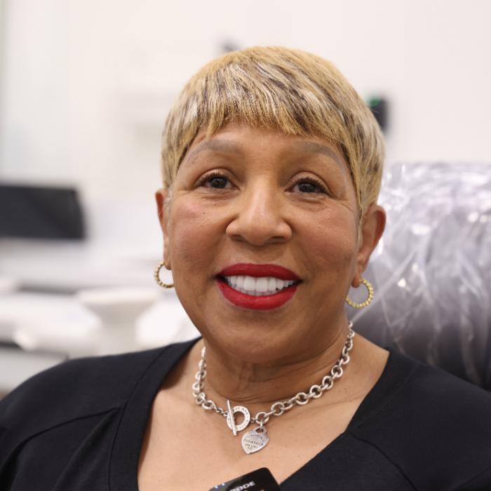 A woman with short blonde hair and red lipstick smiles at the camera, wearing hoop earrings and a silver necklace, seated indoors.