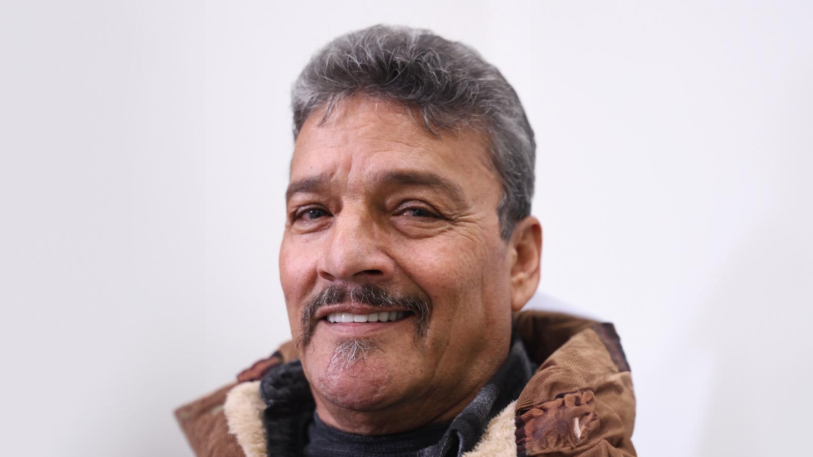 A middle-aged man with gray hair and a mustache, wearing a brown winter jacket, smiles at the camera against a plain white background.