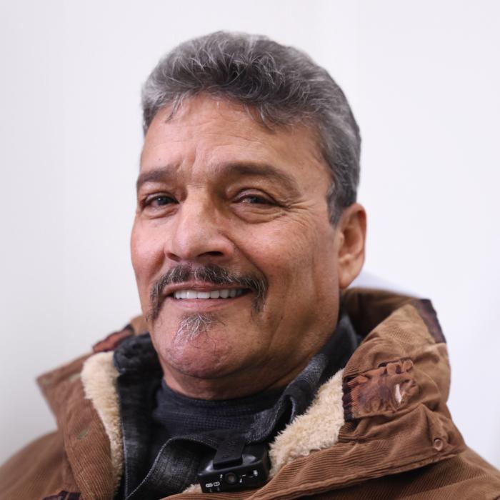 A middle-aged man with gray hair and a mustache, wearing a brown winter jacket, smiling at the camera against a plain white background.