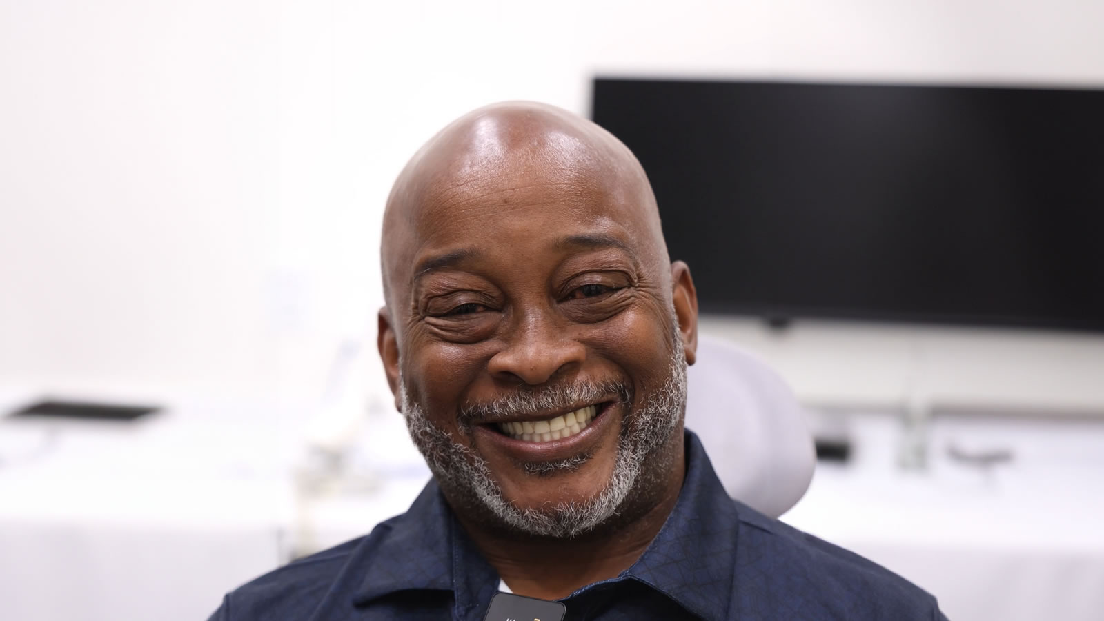 A middle-aged man with a beard smiles while seated indoors in front of a blurred desk and monitor.
