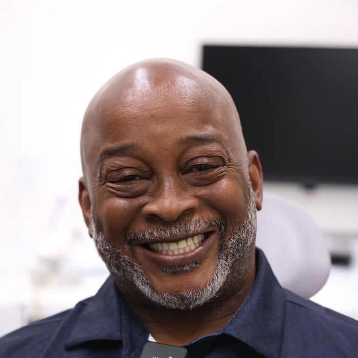 A middle-aged man with a gray beard smiles while seated indoors, with a blurred computer monitor and office equipment in the background.