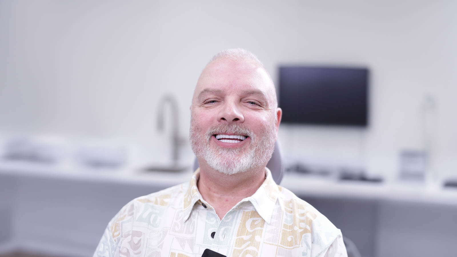 A middle-aged man with a white beard smiling in a modern, bright office setting with a blurred background.