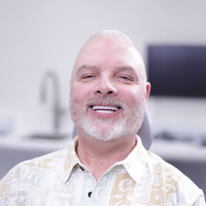 A middle-aged man with light hair and a beard smiles while sitting in a brightly lit room with blurred office equipment in the background.