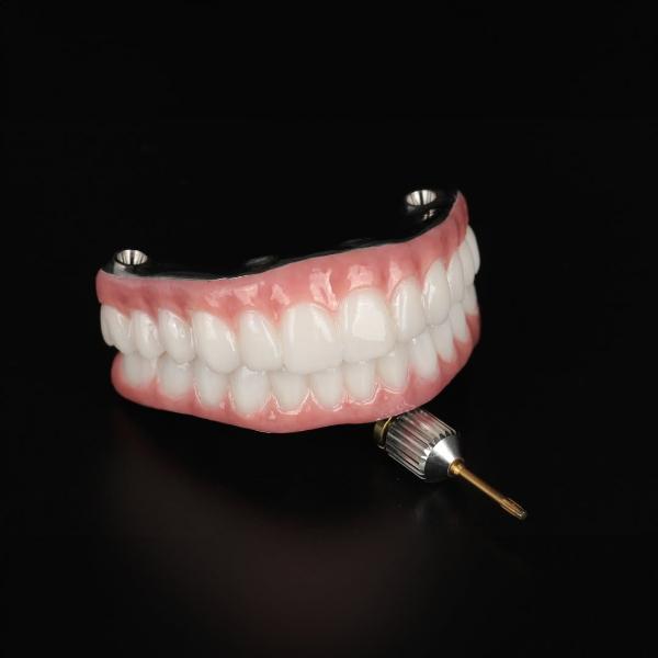 A set of artificial upper and lower teeth with pink gums is displayed on a black background, with a small dental tool placed in front.