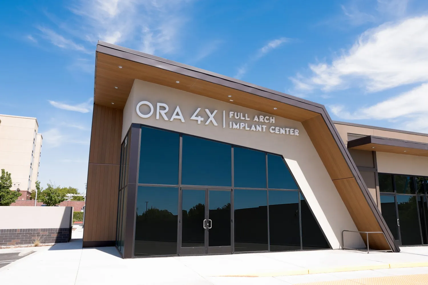Modern building exterior with large glass windows and a sign reading "ORA 4X FULL ARCH IMPLANT CENTER" under a clear blue sky.