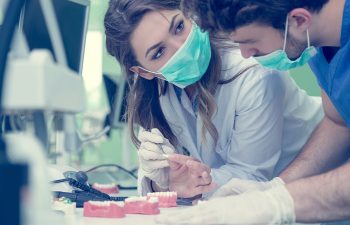 Two dental professionals wearing masks and gloves examine dental models and discuss details at a desk in a clinical setting.