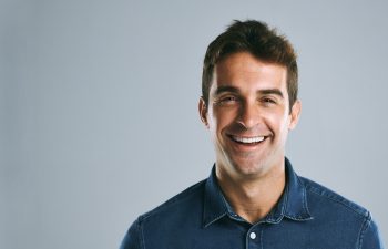 A handsome man with brown hair, wearing a dark blue denim shirt, smiles widely and genuinely while looking directly at the camera. The background is a solid light grey.