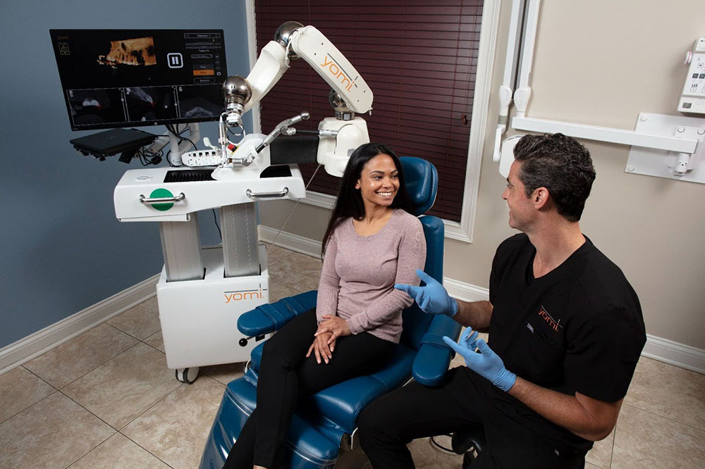 A dentist in scrubs talks to a seated patient next to a robotic dental system with a monitor and robotic arm in a clinic setting.