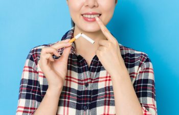 A close-up of a smiling woman in a plaid shirt holding a broken cigarette in one hand and pointing to her white teeth with the other, set against a solid blue background.