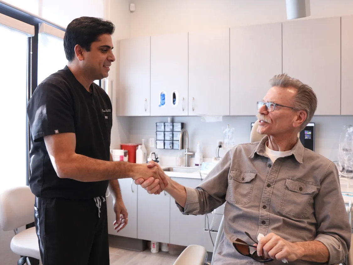 A dentist in scrubs shakes hands with an older male patient seated in a dental clinic, both appearing engaged in conversation.